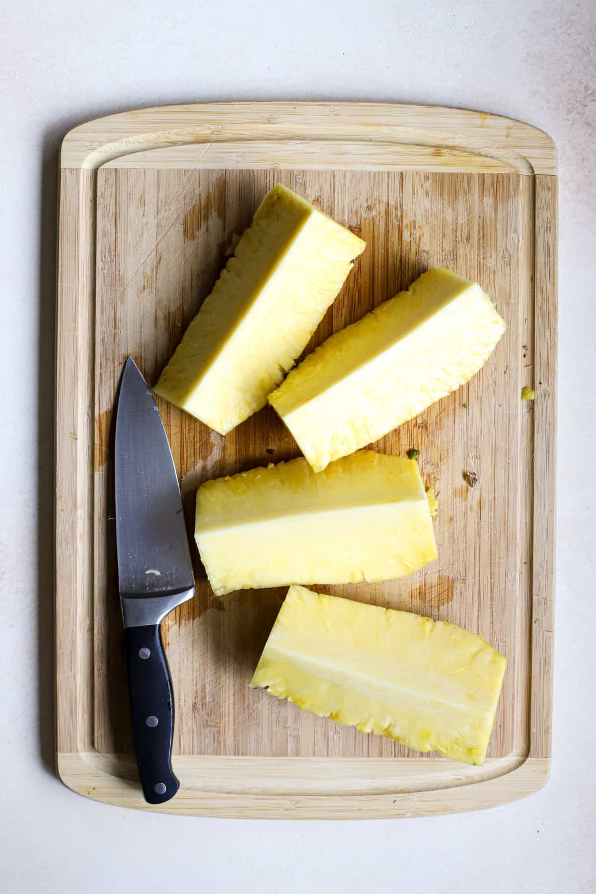 Whole pineapple cut into quarter lengthwise, next to chef's knife on bamboo cutting board