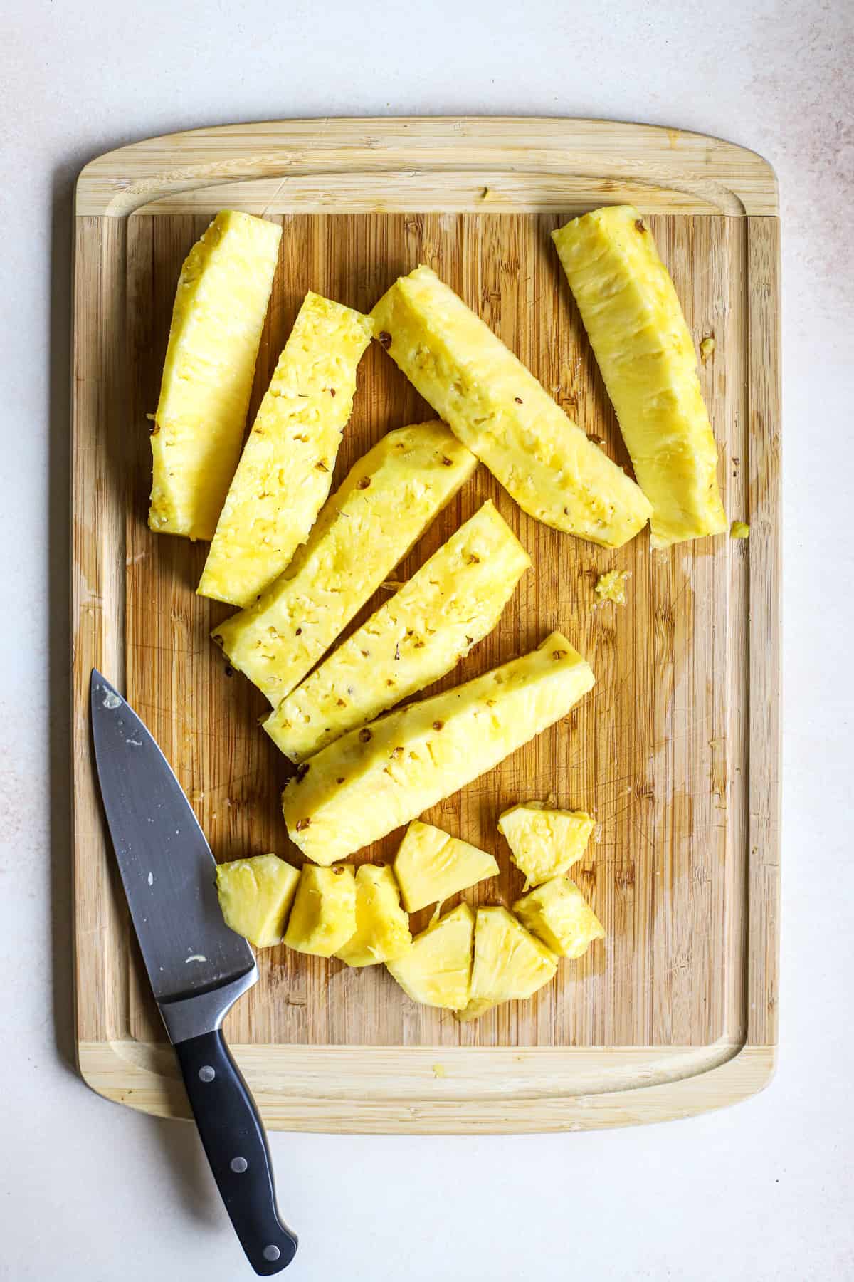 Fresh pineapple sliced lengthwise, with one piece cut into chunks on bamboo cutting board, next to chef's knife