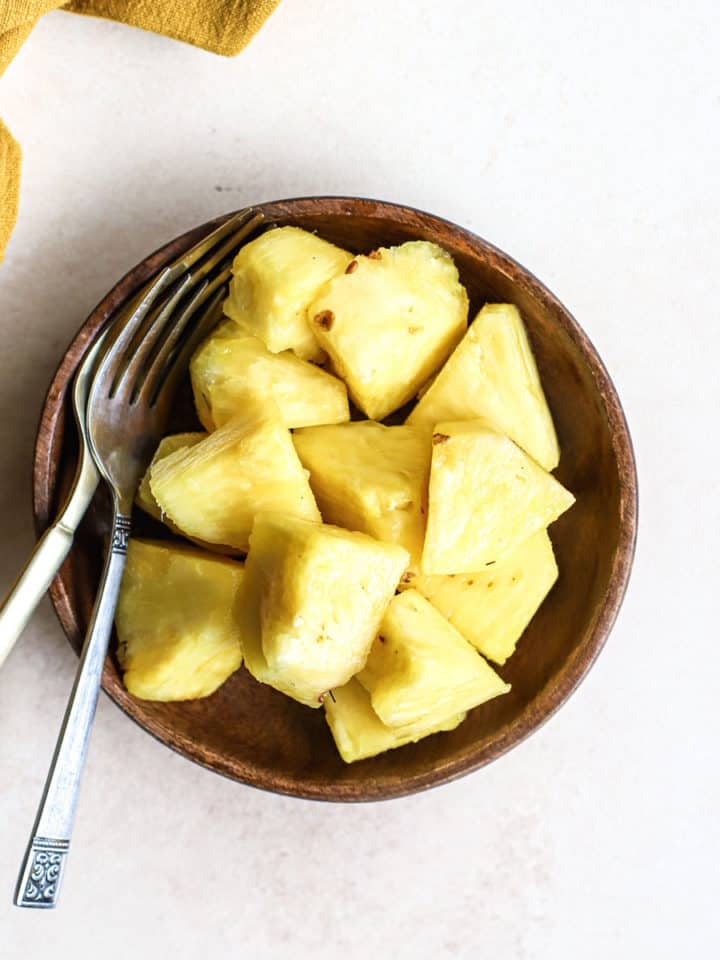 Fresh pineapple chunks on small wooden plate with two forks on beige and white surface, next to gold linen