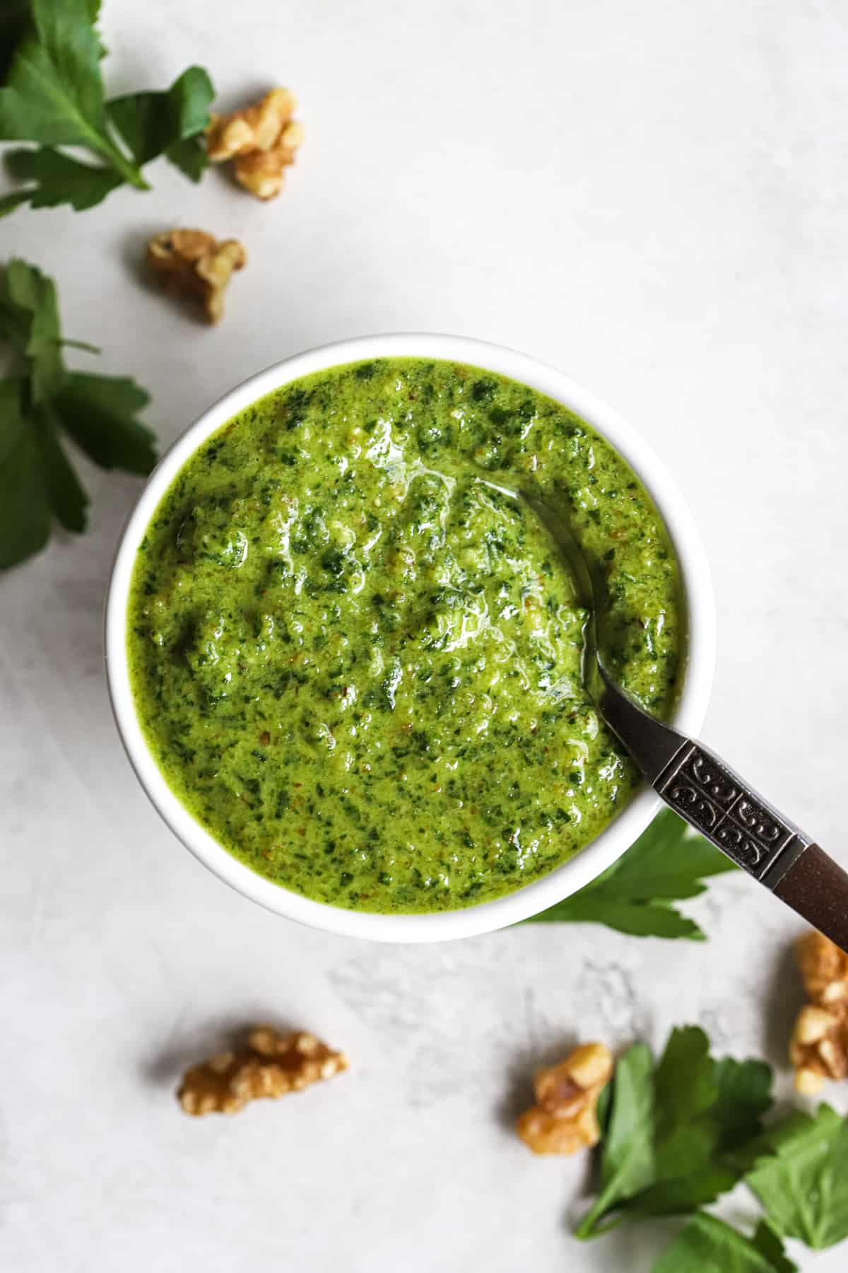 Parsley walnut pesto in small white bowl with spoon, on gray surface with walnuts and parsley leaves sprinkled around