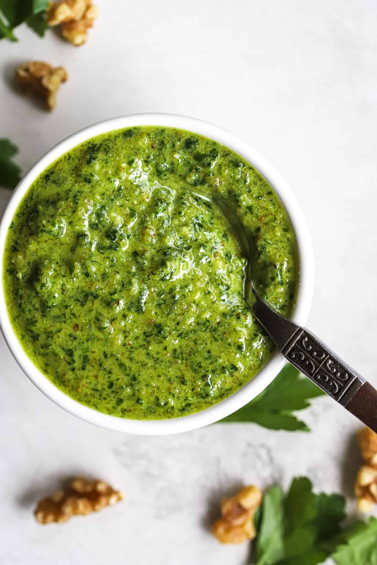 Parsley walnut pesto in small white bowl with spoon, on gray surface with walnuts and parsley leaves sprinkled around