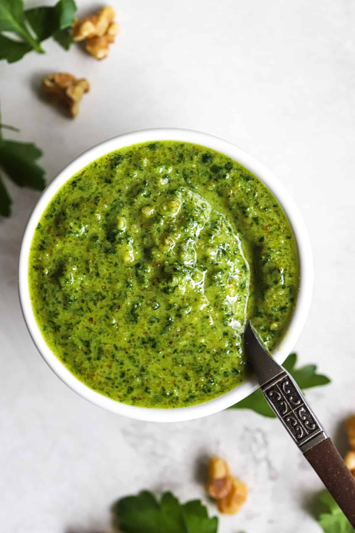 Parsley walnut pesto in small white bowl with spoon, on gray surface with walnuts and parsley leaves sprinkled around