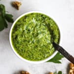Parsley walnut pesto in small white bowl with spoon, on gray surface with walnuts and parsley leaves sprinkled around