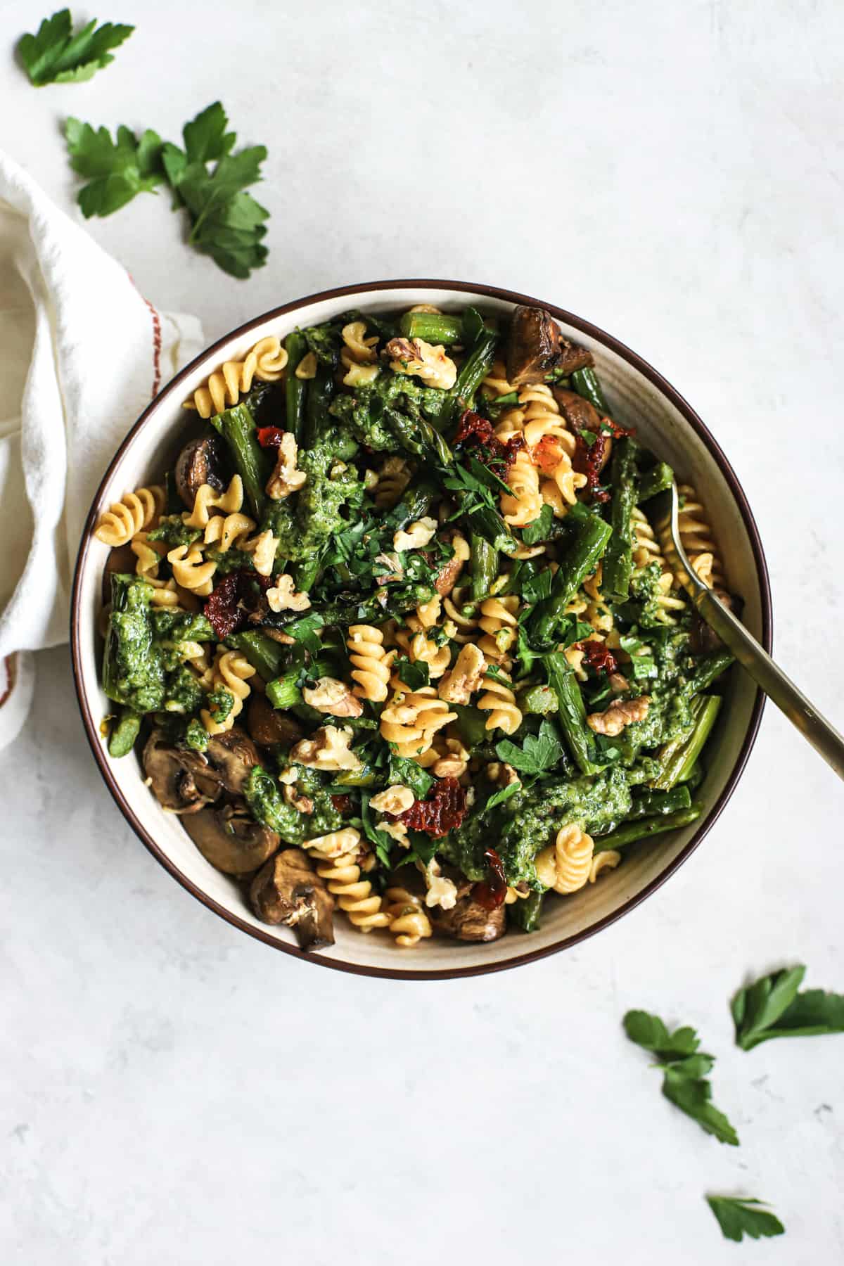 Pesto pasta with spring roasted veggies in beige bowl with golden fork on gray and white surface with parsley sprinkled around