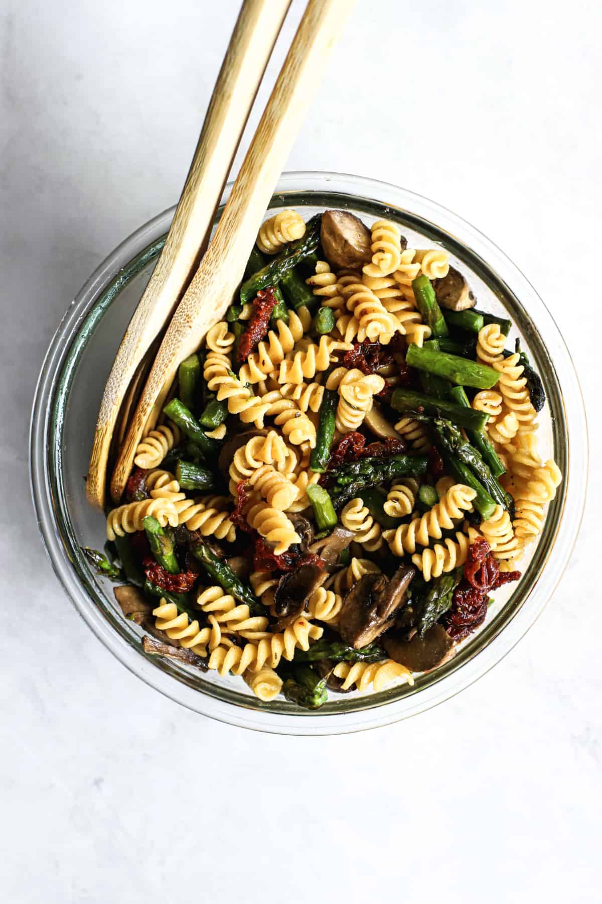 Roasted mushrooms, asparagus, sun-dried tomatoes, and rotini tossed together in clear glass bowl with two wooden spoons on gray/white surface