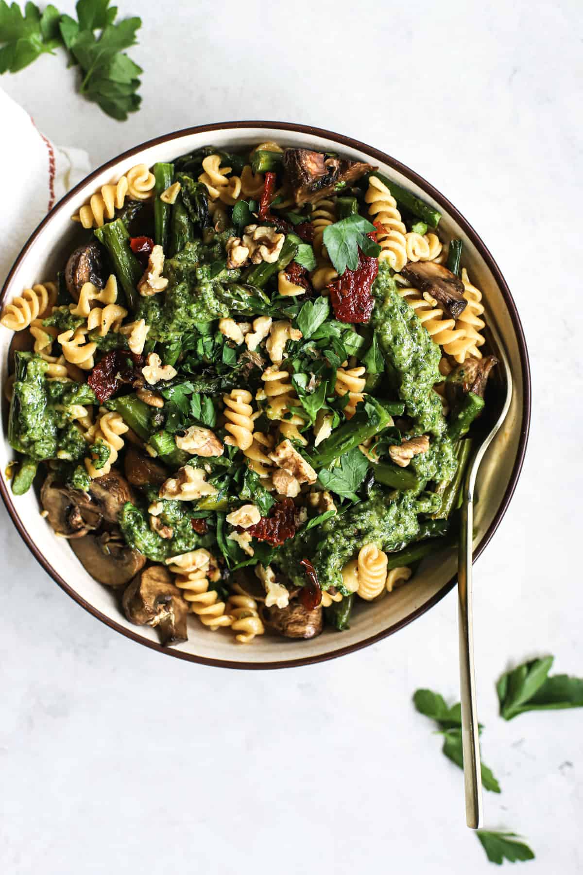 Pesto pasta with spring roasted veggies in beige bowl with golden fork on gray and white surface with parsley sprinkled around