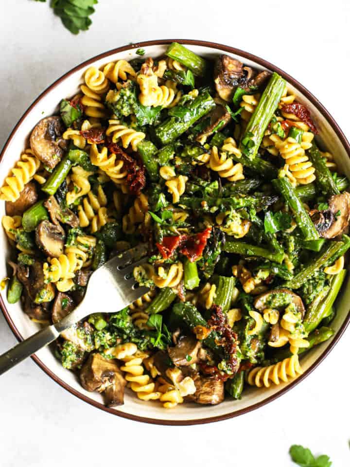 Pesto pasta with spring roasted veggies in beige bowl with golden fork on gray and white surface with parsley sprinkled around