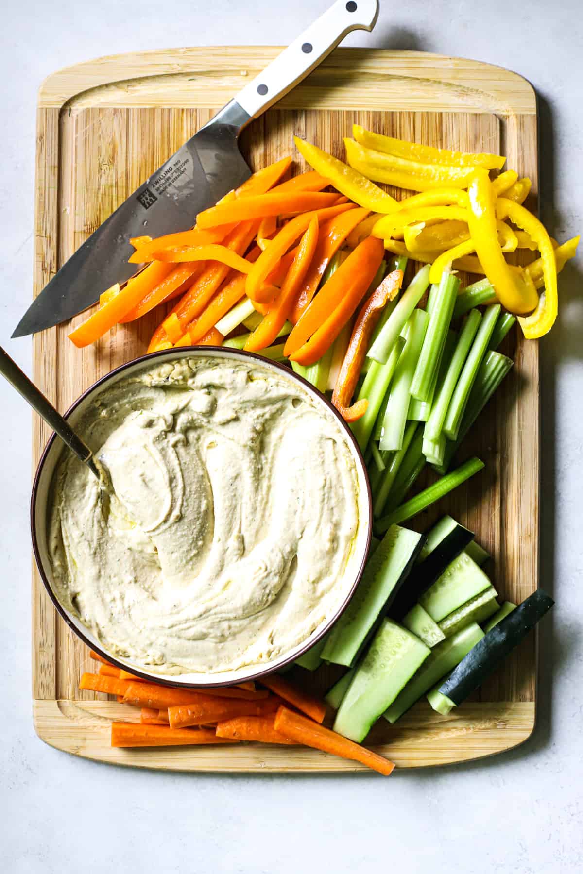 Carrots, cucumbers, celery, and bell peppers sliced on bamboo cutting board next to bowl with hummus and chef's knife