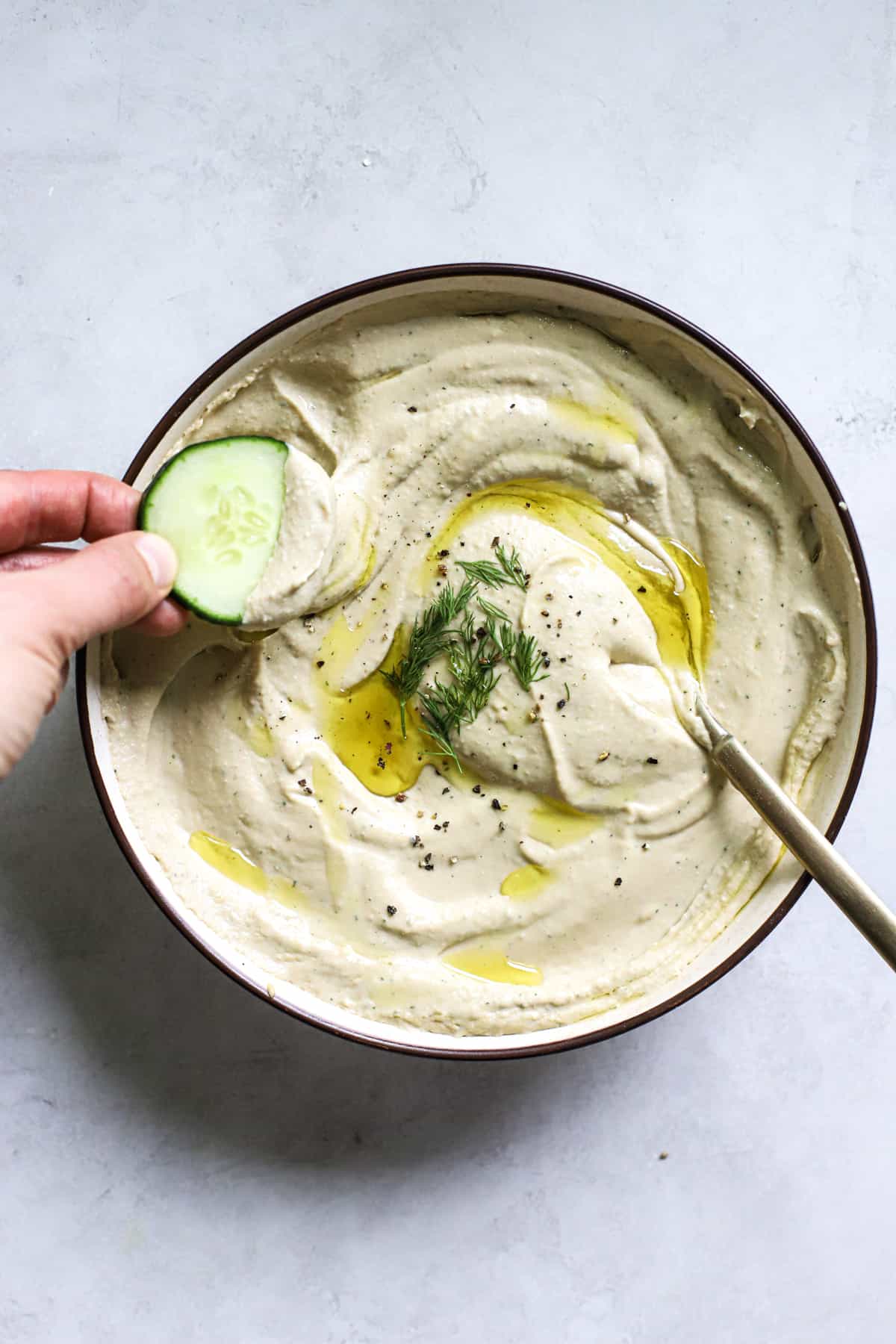 Lemon dill hummus in beige serving dish with olive oil drizzled on top, with golden spoon, and hand dipping a cucumber slice into the hummus.