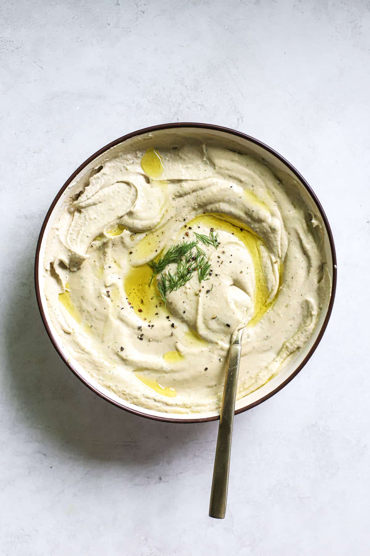 Lemon dill hummus in beige serving dish with olive oil drizzled on top, with golden spoon.