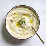 Lemon dill hummus in beige serving dish with olive oil drizzled on top, with golden spoon