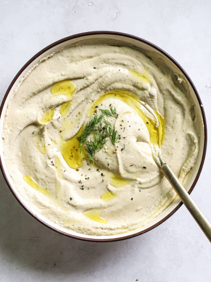 Lemon dill hummus in beige serving dish with olive oil drizzled on top, with golden spoon