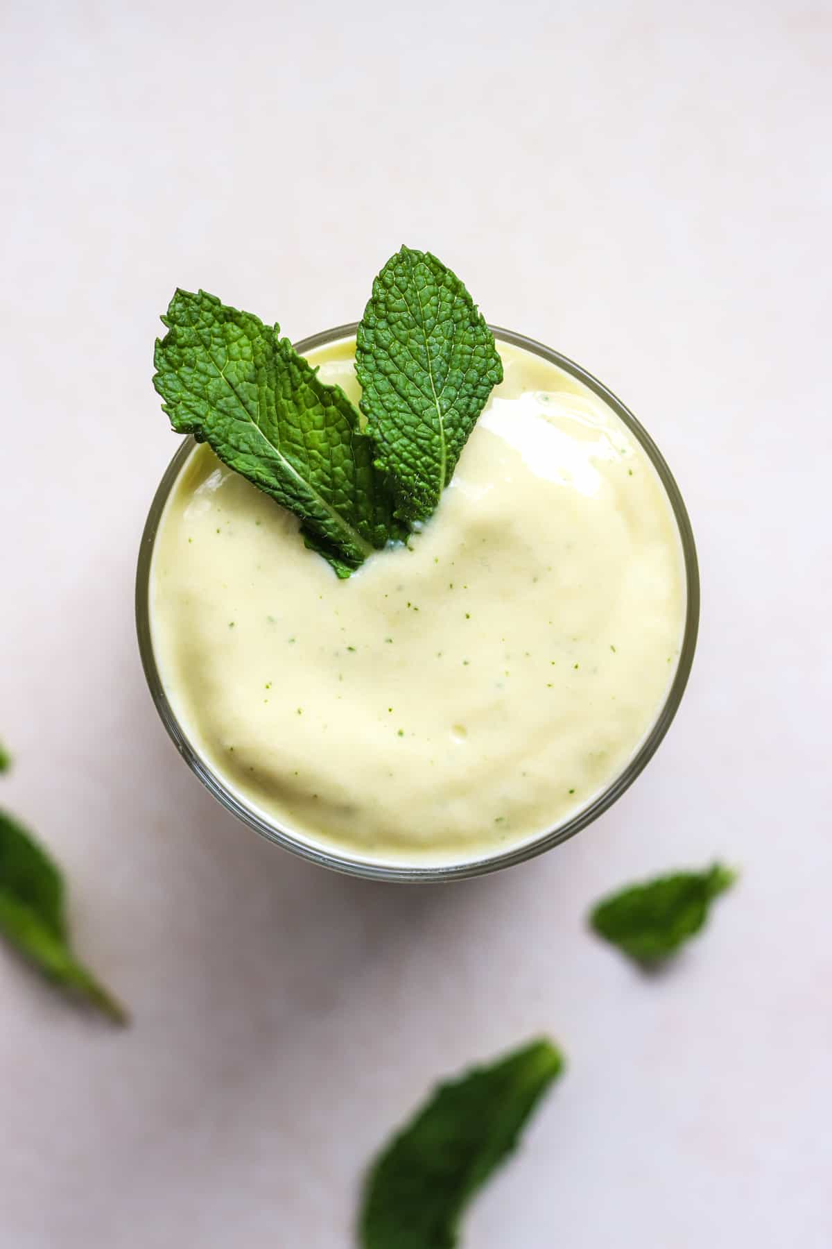 Mango mint smoothie in small glass with fresh mint leaves on top, on light beige and white surface