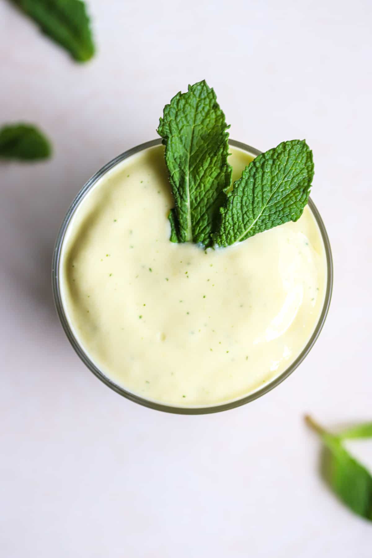 Mango mint smoothie in small glass with fresh mint leaves on top, on light beige and white surface