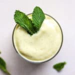 Mango mint smoothie in small glass with fresh mint leaves on top, on light beige and white surface