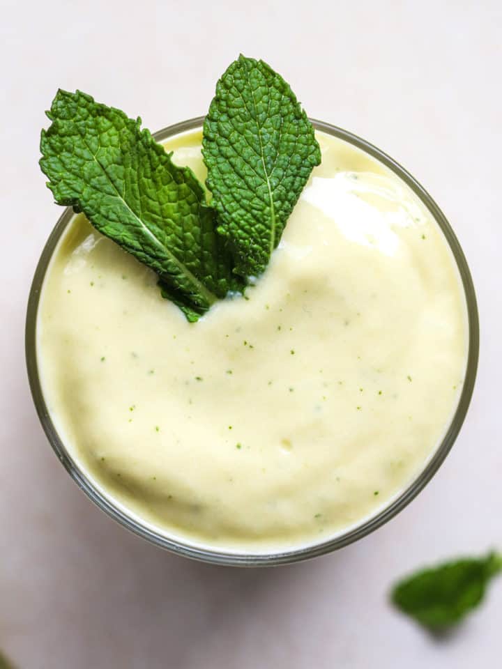 Mango mint smoothie in small glass with fresh mint leaves on top, on light beige and white surface