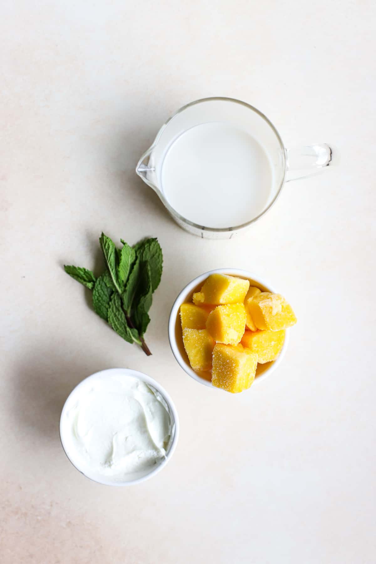 Frozen mango, whole milk Greek yogurt, fresh mint, and milk on beige and white gray surface