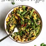 Pesto pasta with spring roasted veggies in beige bowl with golden fork on gray and white surface with parsley sprinkled around