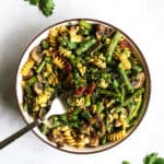 Pesto pasta with spring roasted veggies in beige bowl with golden fork on gray and white surface with parsley sprinkled around