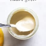 Tahini sauce in small glass jar with spoon and lemons on the side, on gray and white surface