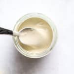 Tahini sauce in small glass jar with spoon and lemons on the side, on gray and white surface