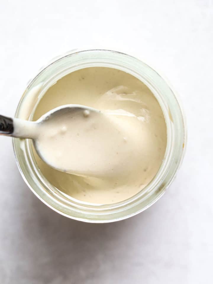 Tahini sauce in small glass jar with spoon and lemons on the side, on gray and white surface