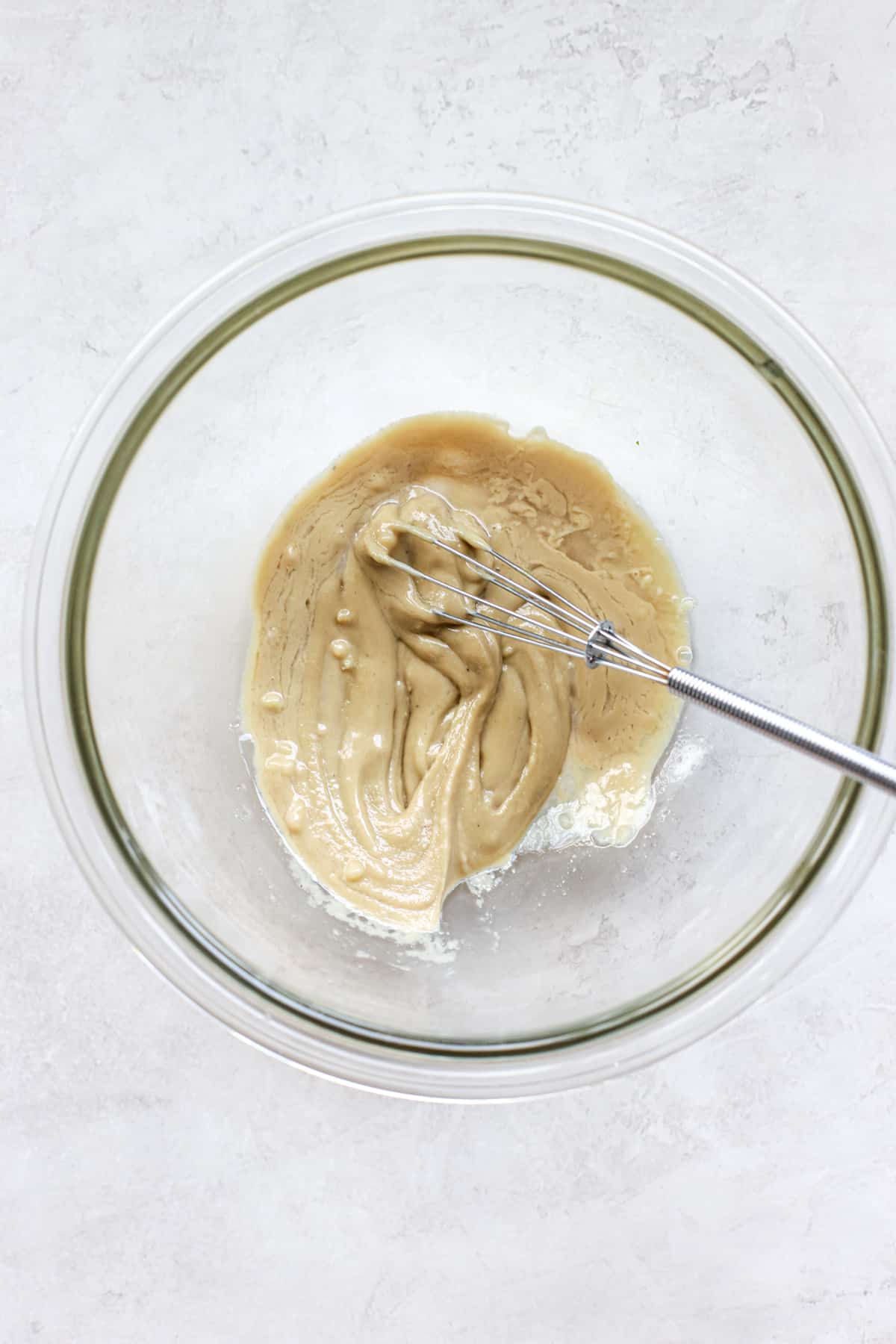 Tahini, lemon juice, garlic, and water in small clear glass bowl with mini whisk, on gray and white surface.