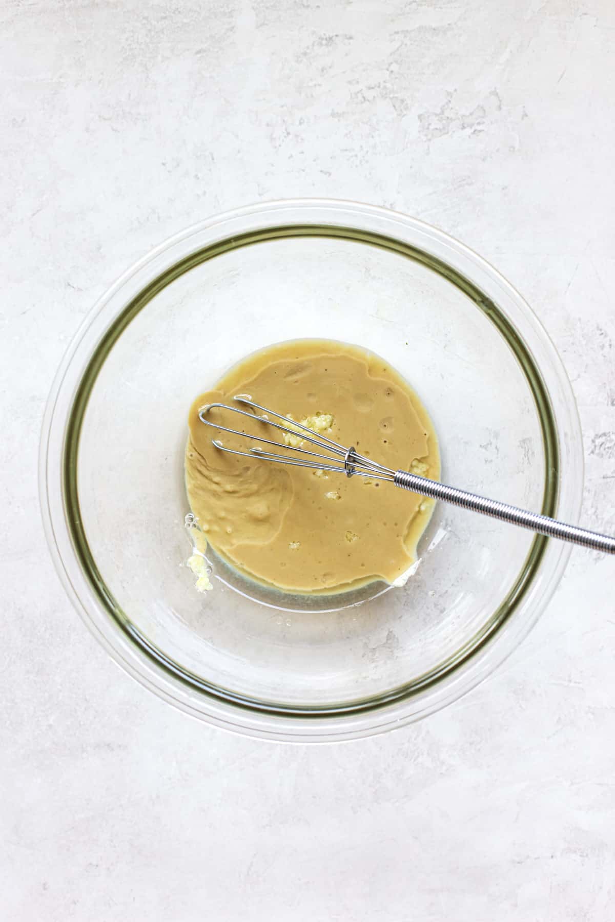 Tahini, lemon juice, and garlic in small clear glass bowl with mini whisk, on gray and white surface.