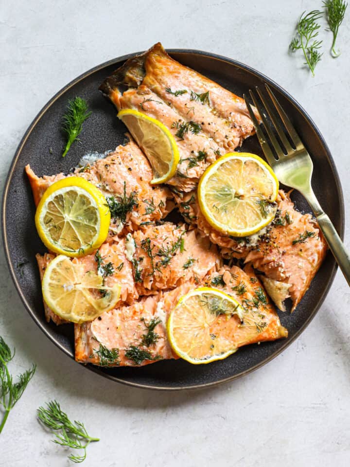 Lemon dill salmon pieces topped with lemon slices, on dark gray plate with fork