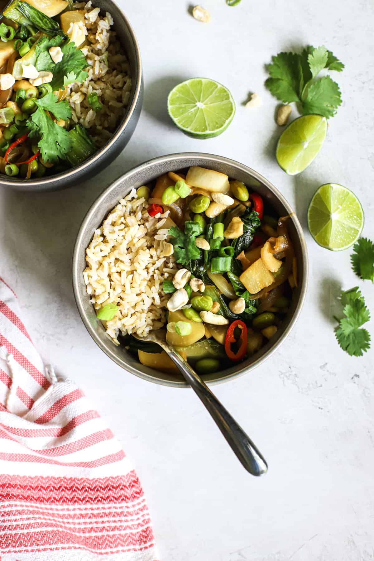 Two gray bowls with pad pak, served with brown rice, with limes wedges and cilantro on the side