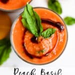 Peach basil gazpacho in clear glass with balsamic glaze swirl on top with basil leaf garnish, on white surface
