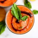 Peach basil gazpacho in clear glass with balsamic glaze swirl on top with basil leaf garnish, on white surface