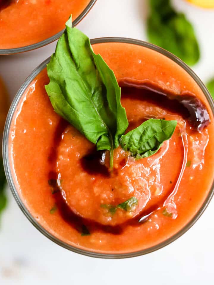 Peach basil gazpacho in clear glass with balsamic glaze swirl on top with basil leaf garnish, on white surface