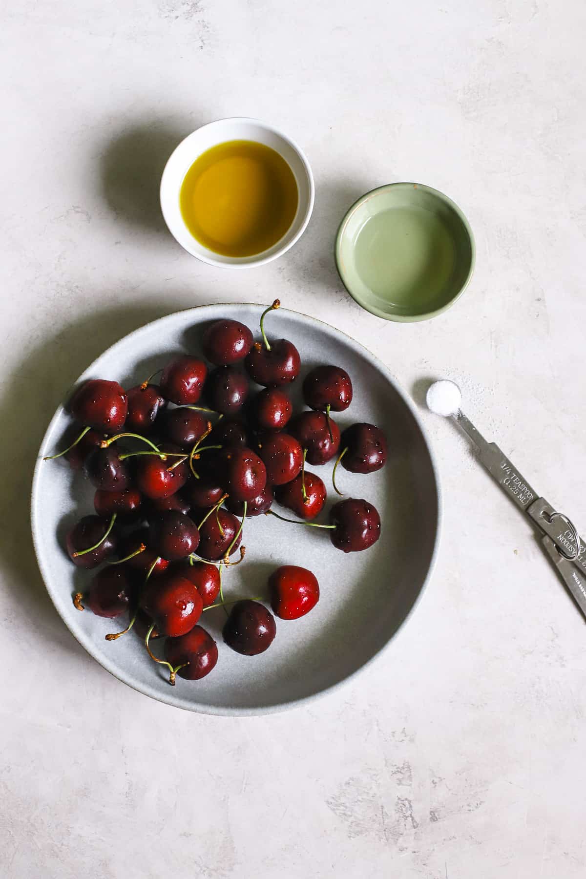 Vinaigrette ingredients: sweet cherries, olive oil, light vinegar, and salt on gray-white surface.