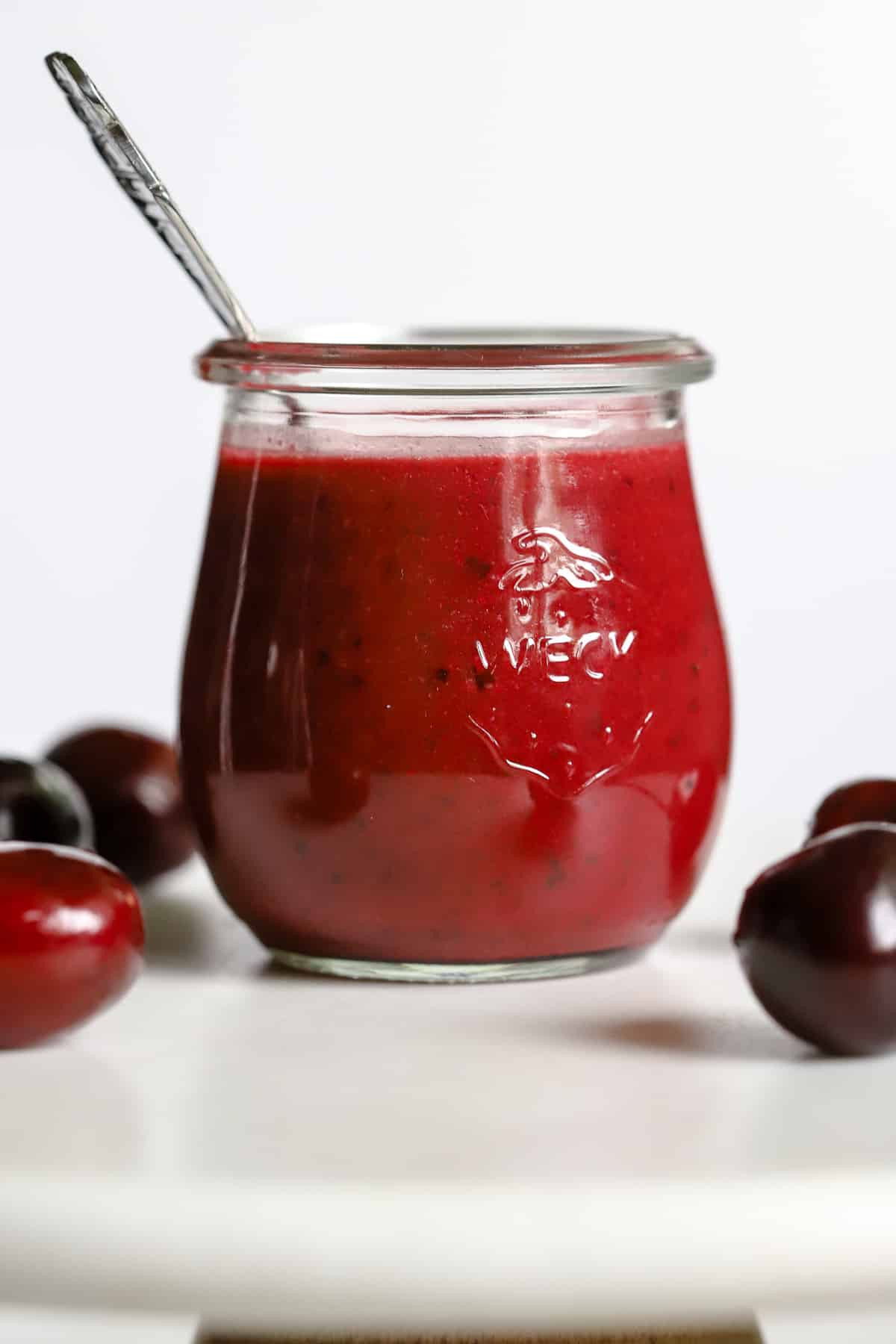 Cherry vinaigrette in small glass jar with spoon, on white marble stand with a few cherries.