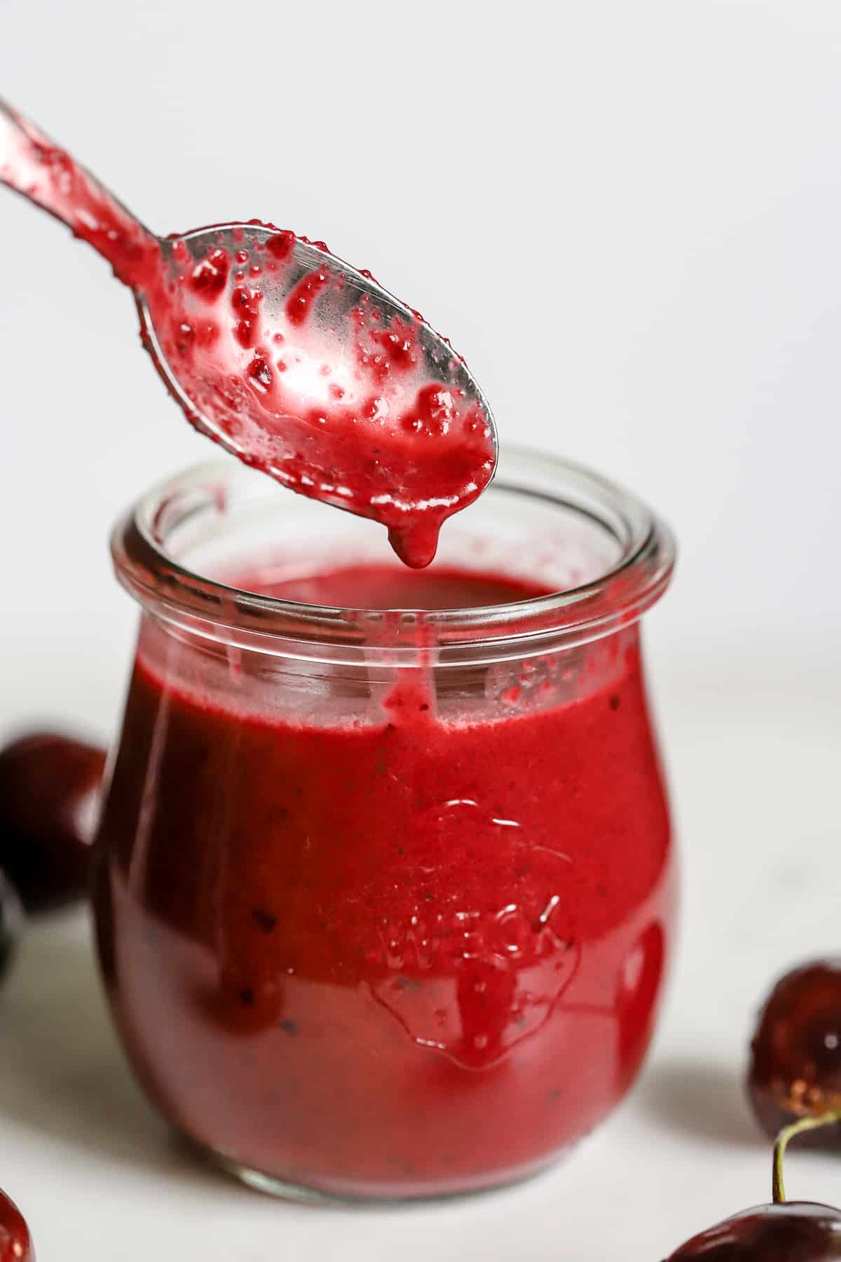 Cherry vinaigrette in small glass jar with spoon dripping with vinaigrette over the top, on white-gray surface with cherries.