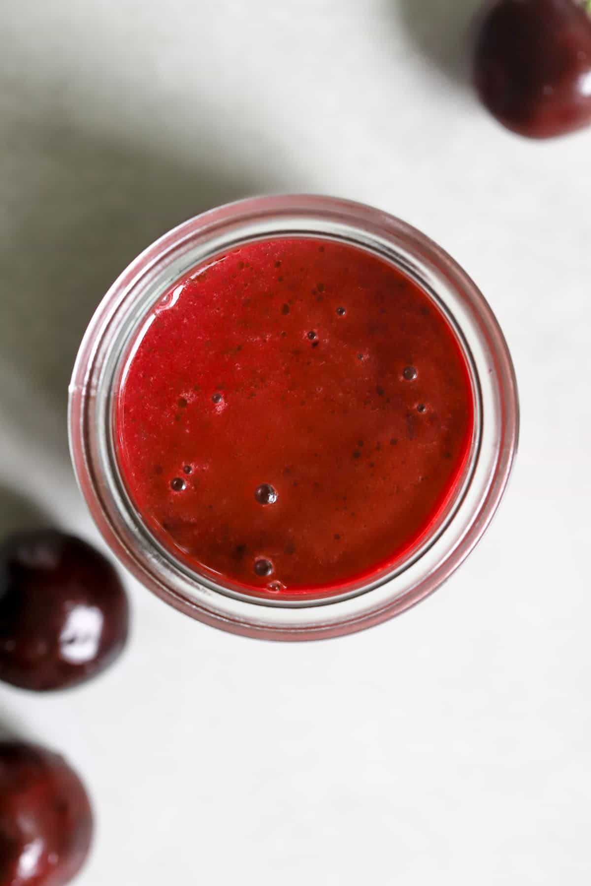 Small glass jar of cherry vinaigrette on gray-white surface with a few cherries.