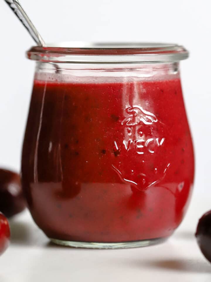 Cherry vinaigrette in small glass jar with spoon, on white marble stand with a few cherries.