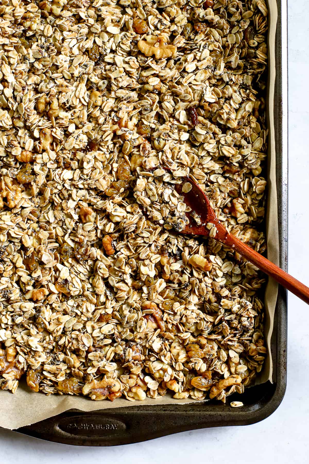 Maple cinnamon walnut granola spread evenly onto parchment-lined cookie sheet with wooden spoon