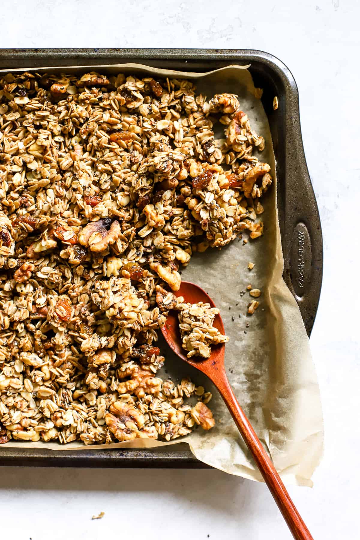 Maple cinnamon walnut granola fully baked, on parchment-lined sheet pan with wooden spoon, and a few clusters broken up