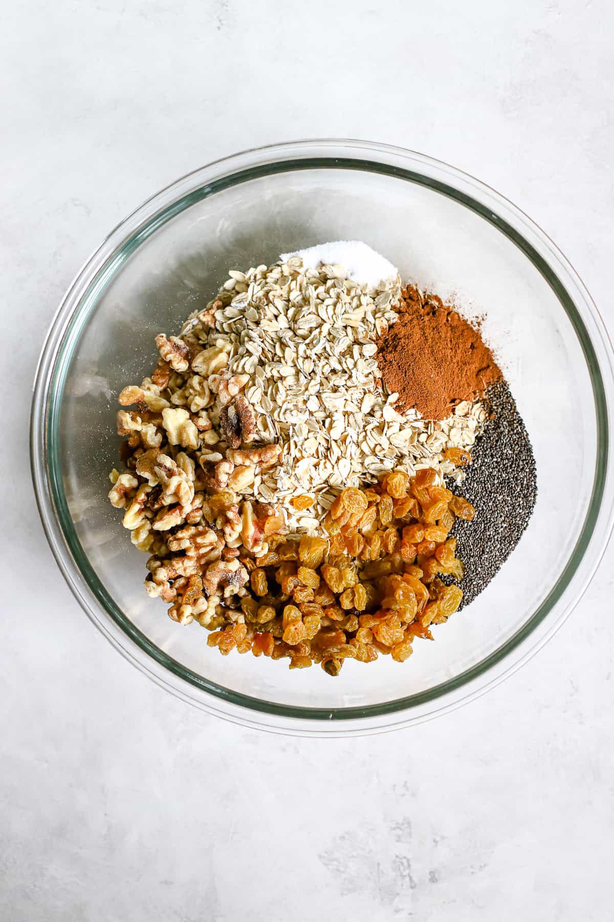 Oats, salt, cinnamon, chia seeds, walnuts, and golden raisins in a clear glass bowl on gray/white surface