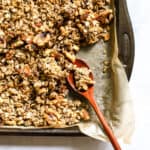 Maple cinnamon walnut granola fully baked, on parchment-lined sheet pan with wooden spoon, and a few clusters broken up