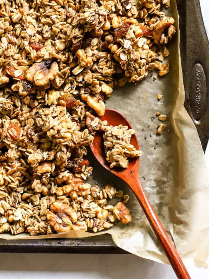 Maple cinnamon walnut granola fully baked, on parchment-lined sheet pan with wooden spoon, and a few clusters broken up
