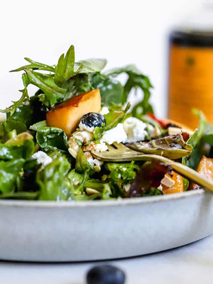 Peach blueberry salad with fork spearing a peach, in light blue bowl with aged balsamic in background