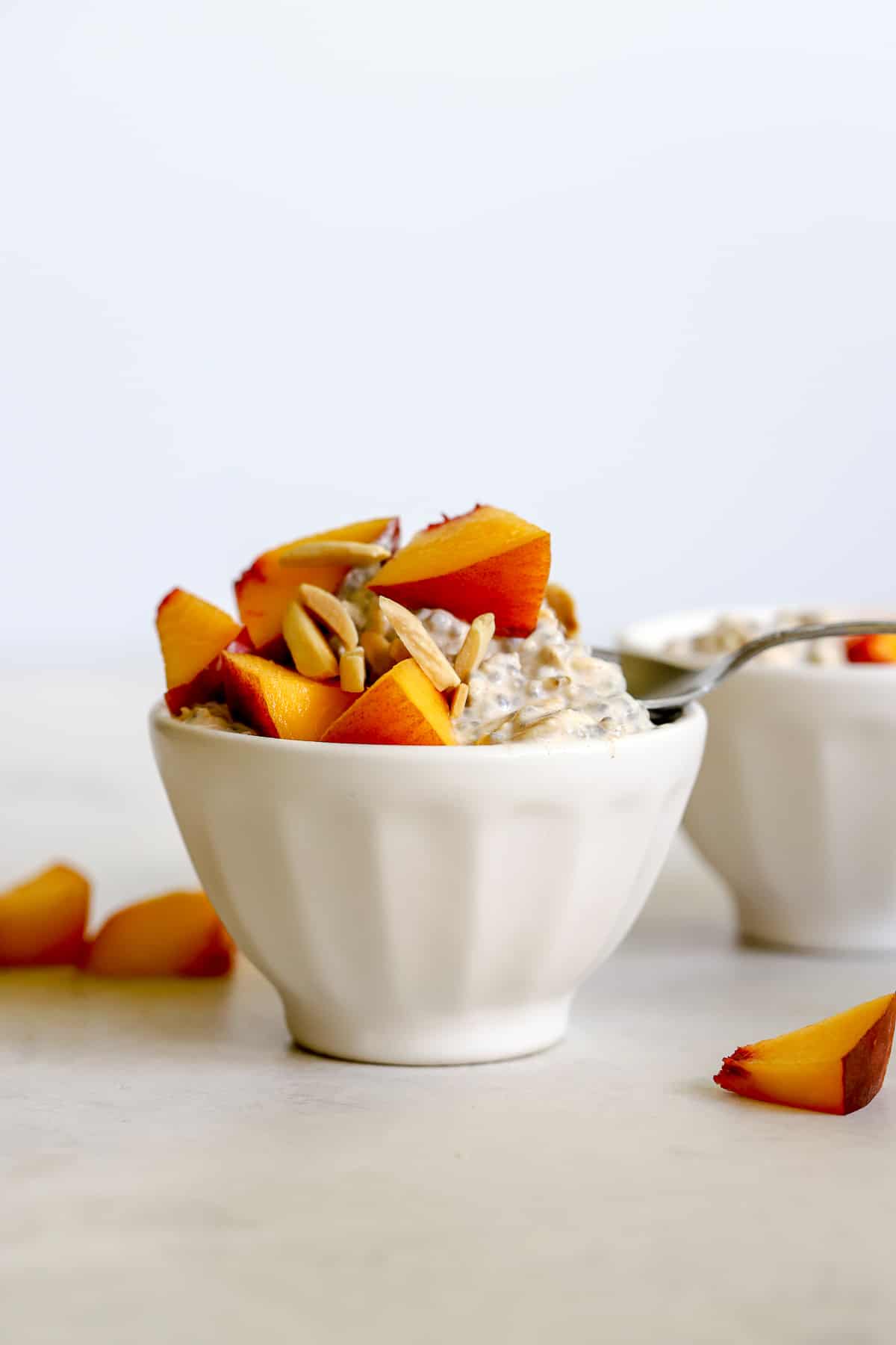 Peach pie overnight oats in small white bowl with diced fresh peaches, slivered almonds, and a dusting of cinnamon, on light gray surface with a few diced peaches on the side