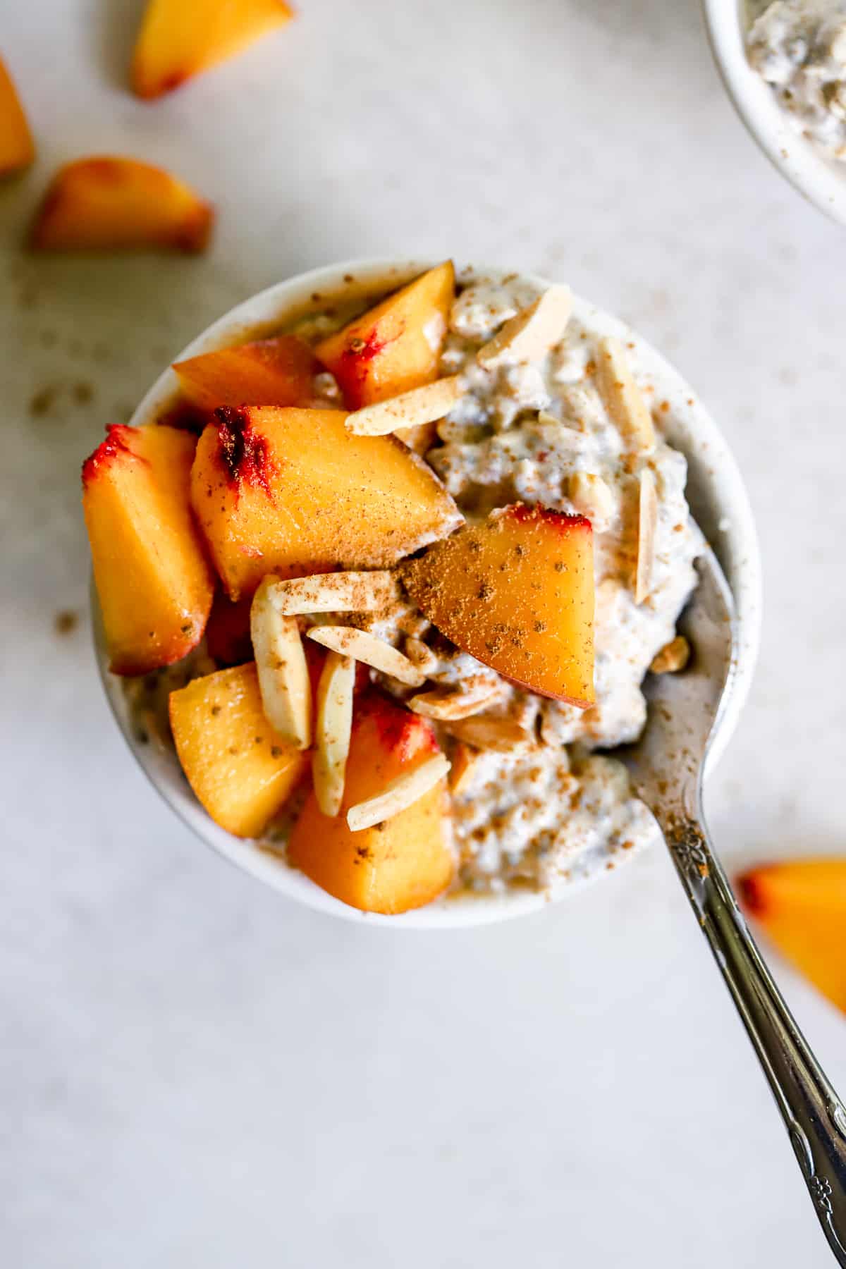 Peach pie overnight oats in small white bowl with diced fresh peaches, slivered almonds, and a dusting of cinnamon