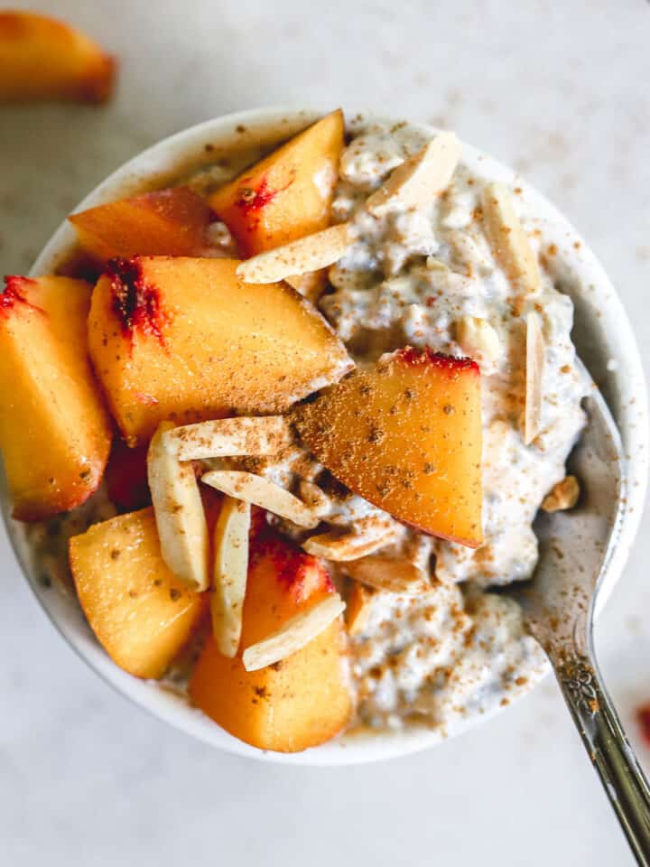 Peach pie overnight oats in small white bowl with diced fresh peaches, slivered almonds, and a dusting of cinnamon