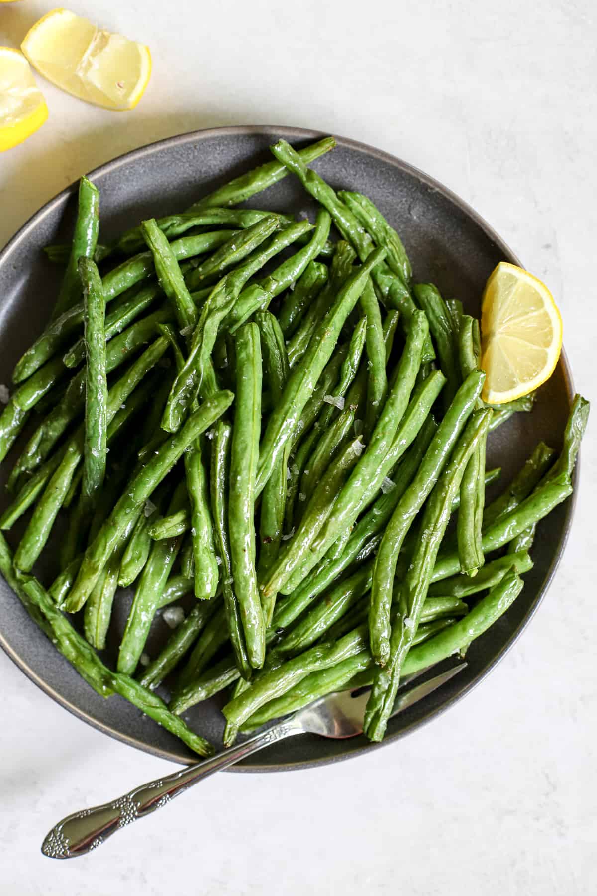 Air fryer green beans on gray plate with lemon wedges, on gray and white surface.