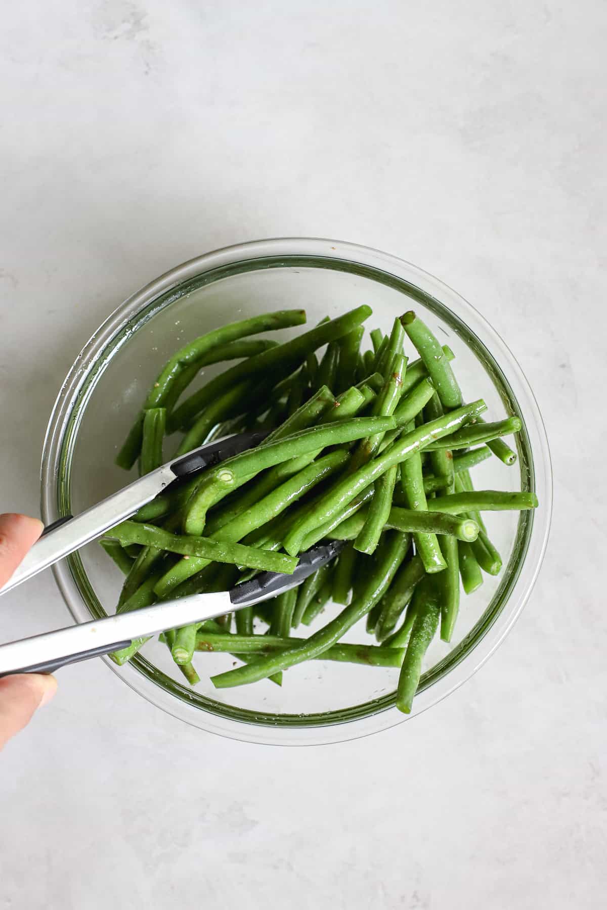 Green beans being tossed in olive oil, salt, and pepper with tongs in clear glass bowl.