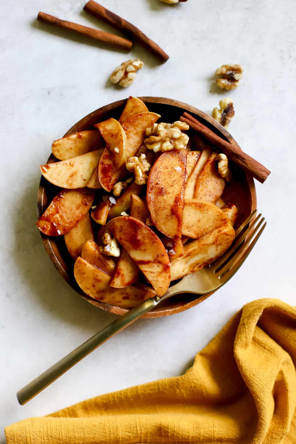 Simple sautéed apples on small wooden plate, topped with walnuts and flaky salt, with a golden fork on gray/white surface with golden yellow linen and cinnamon sticks on side.